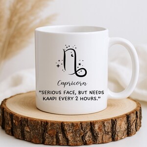 May include: White ceramic mug featuring the Capricorn zodiac symbol and the word "Capricorn" in a stylish font. The mug also includes the quote: "SERIOUS FACE, BUT NEEDS KAAPI EVERY 2 HOURS." The mug is placed on a wooden coaster.