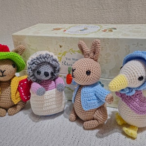 May include: Four crocheted stuffed animals: a rabbit wearing a blue jacket, a hedgehog wearing a pink dress, a duck wearing a purple dress and a yellow beak, and a bunny wearing a yellow jacket and a green hat. The animals are sitting in front of a box with the words "The World of Peter Rabbit" printed on it.