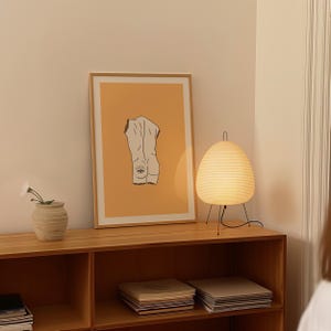 Peut inclure: Une impression encadr&eacute;e d'un dessin au trait d'une figure nue avec un &oelig;il sur le dos, sur fond orange clair. L'impression est expos&eacute;e sur une &eacute;tag&egrave;re en bois avec une lampe blanche et des livres.