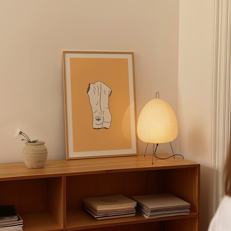 Peut inclure: Une impression encadr&eacute;e d'un dessin au trait d'une figure nue avec un &oelig;il sur le dos, sur fond orange clair. L'impression est expos&eacute;e sur une &eacute;tag&egrave;re en bois avec une lampe blanche et des livres.