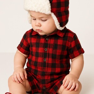 May include: A baby in a red and black check romper suit and matching hat with a white faux fur trim. The baby is also wearing red shoes with laces. The outfit is button-down and the baby is sitting down.