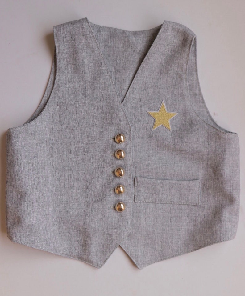 K&ouml;nnte beinhalten: Eine graue Weste mit einem goldenen Stern-Patch und goldenen Kn&ouml;pfen.