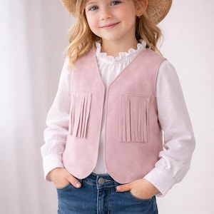 May include: A young child wearing a straw cowboy hat with a pink ribbon and star, a pink suede vest with fringe, a white blouse, and blue jeans. The child is smiling and has their hands in their pockets.