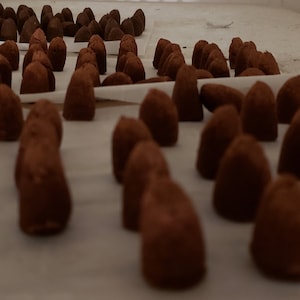 May include: A close-up view of a large quantity of small, dark brown, cone-shaped chocolate truffles arranged on white parchment paper.