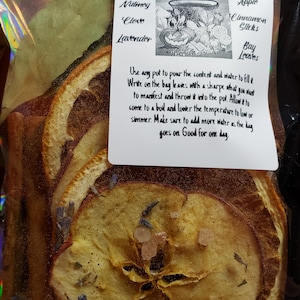 May include: A clear plastic bag filled with dried orange slices, apple slices, cinnamon sticks, bay leaves, and lavender buds. The bag is labeled "Samhain Simmer Pot" and includes instructions for use. The ingredients are for a simmer pot, a type of potpourri that is simmered on the stovetop to create a fragrant aroma.