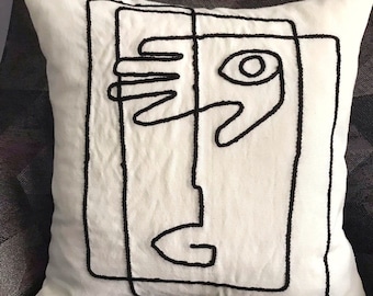 May include: White throw pillow with a black embroidered abstract face design. The face is made up of lines and circles.
