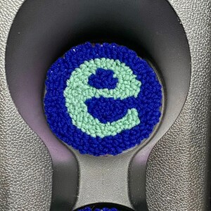 Puede incluir: Dos posavasos circulares azules y verdes con la letra "E" bordada en el centro. Los posavasos están en un portavasos de coche.