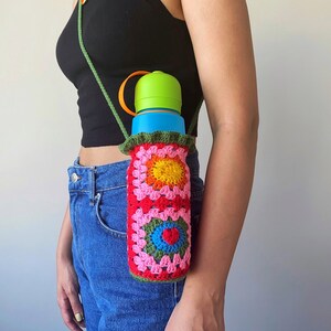 May include: A crocheted water bottle holder with a colorful floral design. The holder is attached to a green strap and worn over the shoulder.