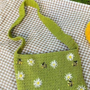 May include: A green crocheted bag with a long strap. The bag is decorated with embroidered white daisies and yellow bees.