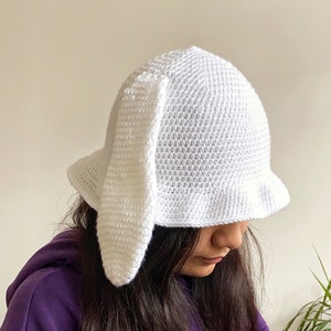 Può includere: Un cappello bianco all'uncinetto con orecchie di coniglio. Il cappello è indossato da una persona con capelli lunghi e scuri e una felpa con cappuccio viola.