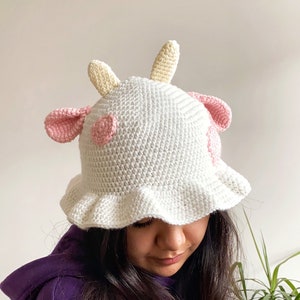 Può includere: Un cappello a secchiello bianco all'uncinetto con dettagli rosa e gialli a forma di mucca. Il cappello ha un'ampia tesa e un bordo arricciato.