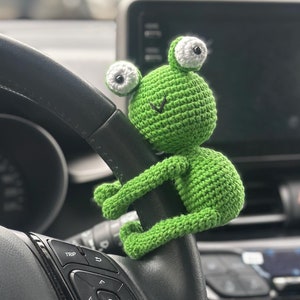 May include: A green crocheted frog toy with white eyes is hanging from the steering wheel of a car. The steering wheel has black leather and buttons for controlling the car's functions.