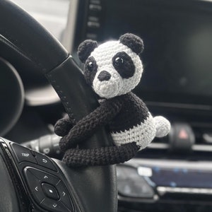 May include: A black and white crocheted panda bear plush toy hanging from a car's steering wheel. The steering wheel has buttons for controlling the car's functions.