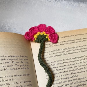 May include: A close-up of a green crocheted bookmark with a pink and yellow flower on top. The bookmark is resting on an open book with text about a woman named Anna.
