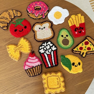 Puede incluir: Un conjunto de posavasos hechos a mano con temática de comida. El conjunto incluye un cruasán, una rosquilla, un huevo frito, un tomate, pan, aguacate, patatas fritas, pasta de lazo, una magdalena, palomitas de maíz, una porción de pizza, un limón y un ravioli.