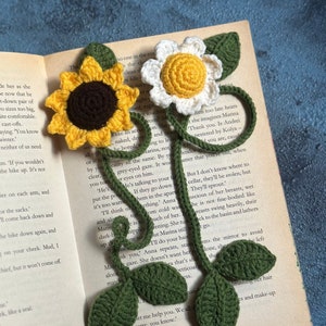 May include: A crocheted bookmark featuring a sunflower, a daisy, and green leaves. The bookmark is placed on an open book with text.