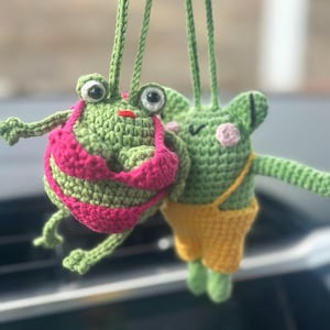 May include: Two handmade crochet frog ornaments. One frog wears a pink and green striped bikini, the other wears yellow overalls. Both frogs are green with large eyes and are suspended by green cords.