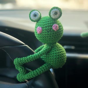 May include: A green crocheted frog ornament with large, round eyes and pink cheeks. The frog is perched on a black steering wheel. The frog's arms and legs are wrapped around the steering wheel.
