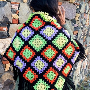 May include: A colorful, crocheted shawl featuring a geometric pattern of squares in black, orange, green, yellow, and purple. The shawl has a fringe trim and is worn over the shoulders.