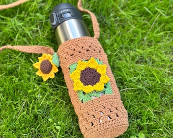 May include: A brown crocheted water bottle holder with a sunflower design. The holder has a strap and is sitting on green grass.