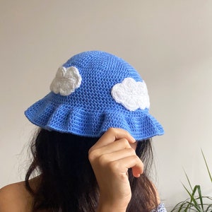 Può includere: Un cappello a secchiello in crochet blu con nuvole bianche in crochet sulla corona. Il cappello ha un bordo arricciato.
