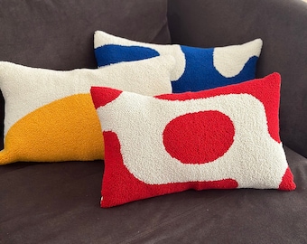 May include: Three decorative throw pillows with abstract designs. The pillows are white with colorful geometric shapes. The pillows are on a dark brown couch.