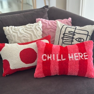 Puede incluir: Una colección de cojines decorativos con varios diseños y colores. Los cojines incluyen blanco con lunares rojos, rosa con un patrón ondulado y un cojín a rayas rojas y rosas con el texto "CHILL HERE". Otros cojines tienen diseños abstractos.