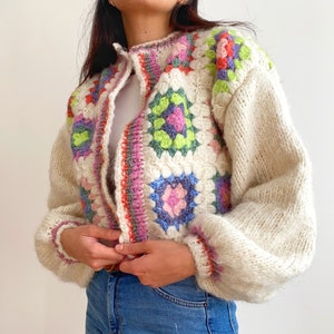 May include: A white cardigan with a crocheted front panel featuring colorful granny squares. The cardigan has a button closure and long sleeves.