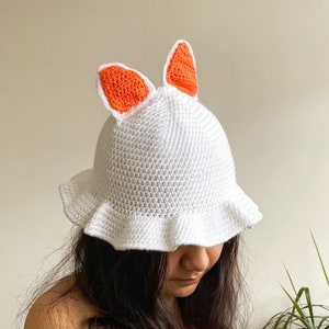 Può includere: Un cappello a secchiello bianco all'uncinetto con orecchie di coniglio arancioni. Il cappello ha un bordo arricciato.