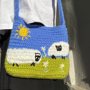 May include: A blue crocheted shoulder bag with a yellow sun, green grass, and two white sheep with black faces. The bag has a long strap.