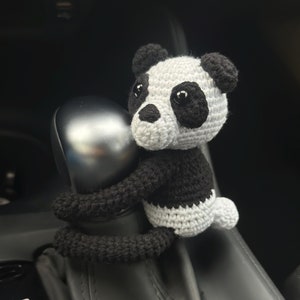 May include: A black and white crocheted panda bear toy is attached to a car's gear stick. The panda has black eyes and a black nose.