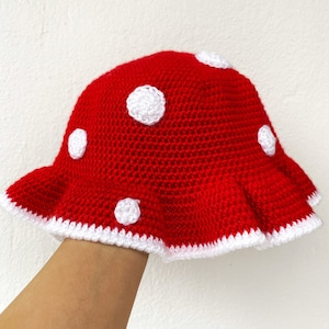 Può includere: Un cappello a forma di fungo di colore rosso lavorato a uncinetto con pois bianchi e un bordo bianco lavorato a uncinetto.