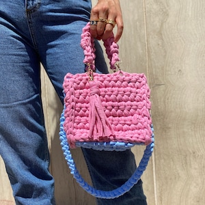 May include: A pink and light blue crocheted handbag with a tassel. The bag has a short handle and a long strap.