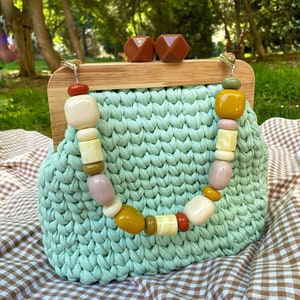 May include: A light green crocheted clutch bag with a wooden frame and a clasp. The bag is decorated with colorful beads in shades of brown, yellow, white, and pink.