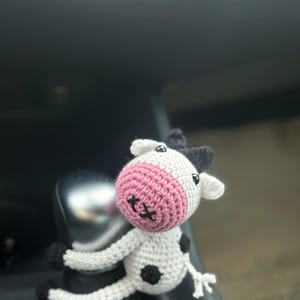 Könnte beinhalten: Eine gehäkelte weiße Kuh mit schwarzen Flecken und rosa Ohren. Die Kuh sitzt auf dem Schalthebel eines Autos.