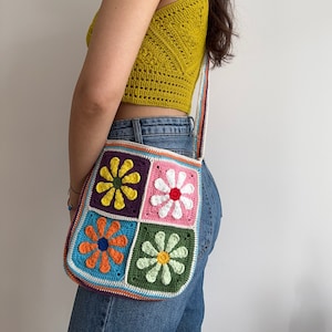 May include: A handmade crochet shoulder bag with a colorful daisy pattern. The bag features a square design with four daisy squares in orange, pink, yellow, and white. The strap is striped with multiple colors. A yellow crochet top is also visible.