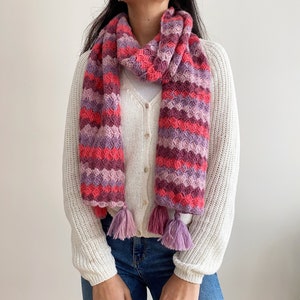 Peut inclure: Une écharpe en crochet rayée rose et violette avec des pompons. L'écharpe est portée par une personne portant un pull blanc et un jean bleu.