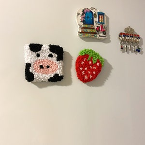 Könnte beinhalten: Eine Sammlung dekorativer Magnete auf einer weißen Oberfläche. Die Magnete zeigen eine Kuh, eine Erdbeere, ein Souvenir mit Schlüsseln und eine farbenfrohe Türszene mit einem Roller. Der Kuhmagnet ist weiß mit schwarzen Flecken und einer rosa Nase.