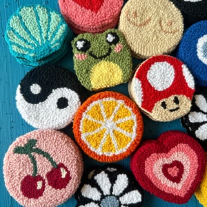 May include: A collection of colorful, round, hand-stitched felt ornaments. The ornaments feature various designs, including a heart, a frog, a mushroom, a daisy, a lemon, a cherry, and a yin-yang symbol.