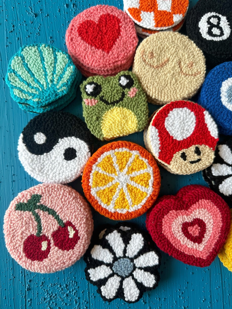 May include: A collection of colorful, round, hand-stitched felt ornaments. The ornaments feature various designs, including a heart, a frog, a mushroom, a daisy, a lemon, a cherry, and a yin-yang symbol.