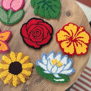 May include: A collection of colorful, handmade floral coasters. The set includes a pink tulip, a green leaf, a red rose, a yellow hibiscus, a sunflower, and a blue and white water lily. The coasters are arranged on a wooden surface.