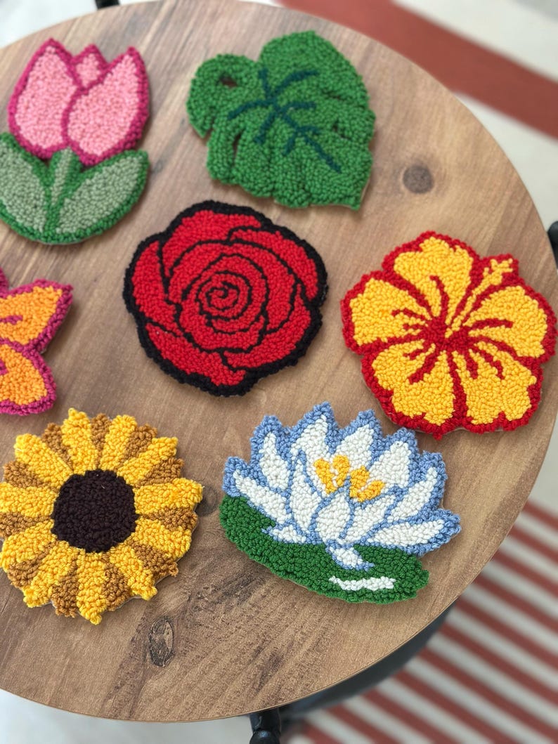 May include: A collection of colorful, handmade floral coasters. The set includes a pink tulip, a green leaf, a red rose, a yellow hibiscus, a sunflower, and a blue and white water lily. The coasters are arranged on a wooden surface.