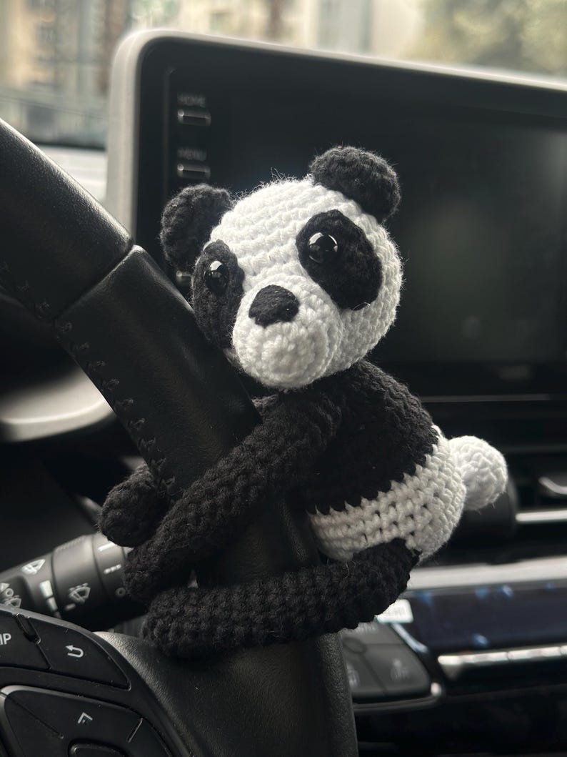 以下が含まれることがあります： 黒と白の毛糸で編まれたパンダのぬいぐるみが車のハンドルにぶら下がっている