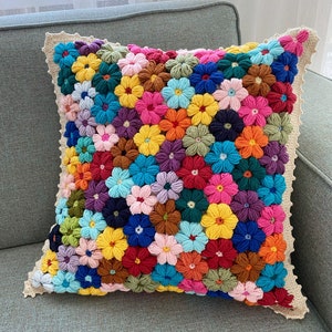 Puede incluir: Una almohada de crochet colorida con un diseño floral. La almohada está hecha de una variedad de colores, incluyendo rosa, azul, amarillo, verde y rojo. La almohada está sentada en un sofá gris.