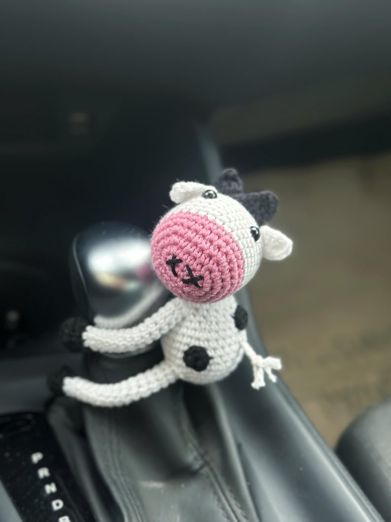 K&ouml;nnte beinhalten: Eine geh&auml;kelte wei&szlig;e Kuh mit schwarzen Flecken und rosa Ohren. Die Kuh sitzt auf dem Schalthebel eines Autos.