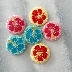 May include: Six round, colorful, felt-covered coasters with embroidered hibiscus flowers. The coasters are in shades of pink, yellow, blue, and green.