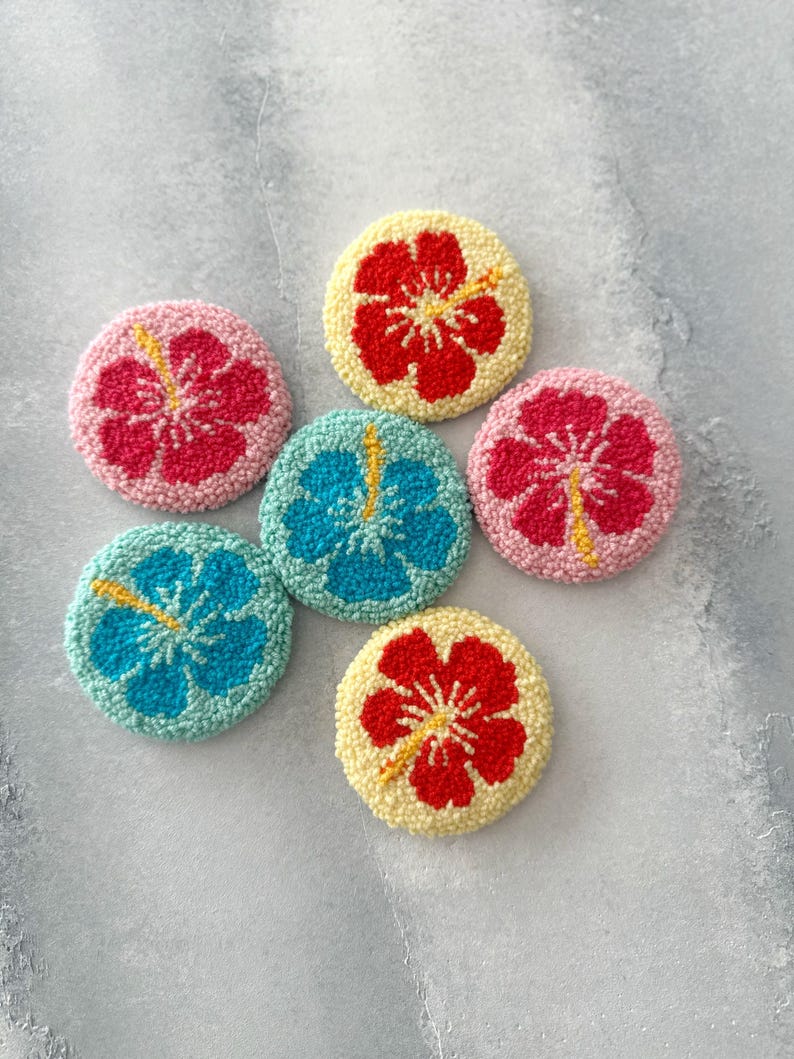 May include: Six round, colorful, felt-covered coasters with embroidered hibiscus flowers. The coasters are in shades of pink, yellow, blue, and green.