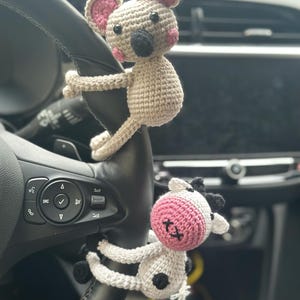 K&ouml;nnte beinhalten: Zwei geh&auml;kelte Stofftiere, eine Maus und eine Kuh, h&auml;ngen am Lenkrad eines Autos. Die Maus ist beige mit rosa Ohren und einer schwarzen Nase. Die Kuh ist wei&szlig; mit schwarzen Flecken und rosa Ohren.