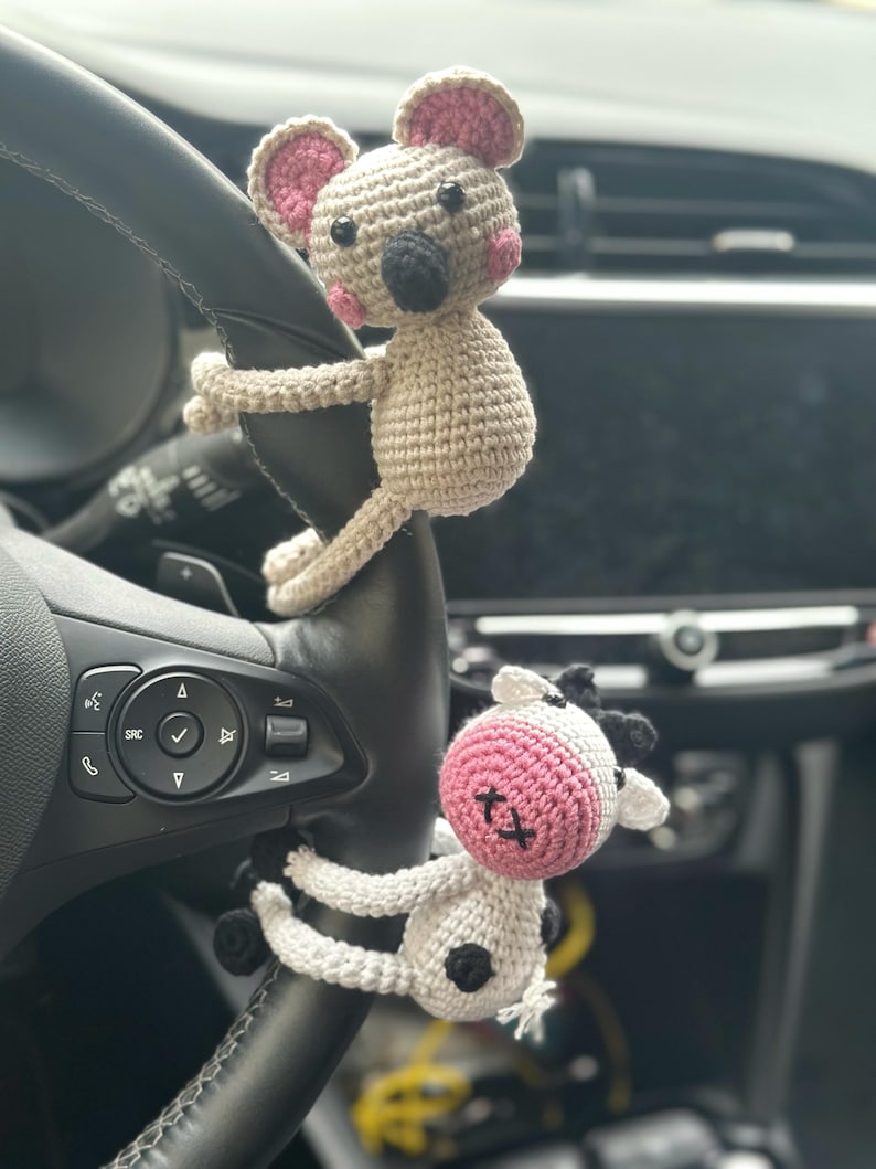 K&ouml;nnte beinhalten: Zwei geh&auml;kelte Stofftiere, eine Maus und eine Kuh, h&auml;ngen am Lenkrad eines Autos. Die Maus ist beige mit rosa Ohren und einer schwarzen Nase. Die Kuh ist wei&szlig; mit schwarzen Flecken und rosa Ohren.