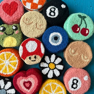 May include: A collection of 12 colorful, round, crocheted coasters with various designs, including a heart, a mushroom, a daisy, a lemon, a cherry, an eye, a frog, a checkered pattern, and the number 8.